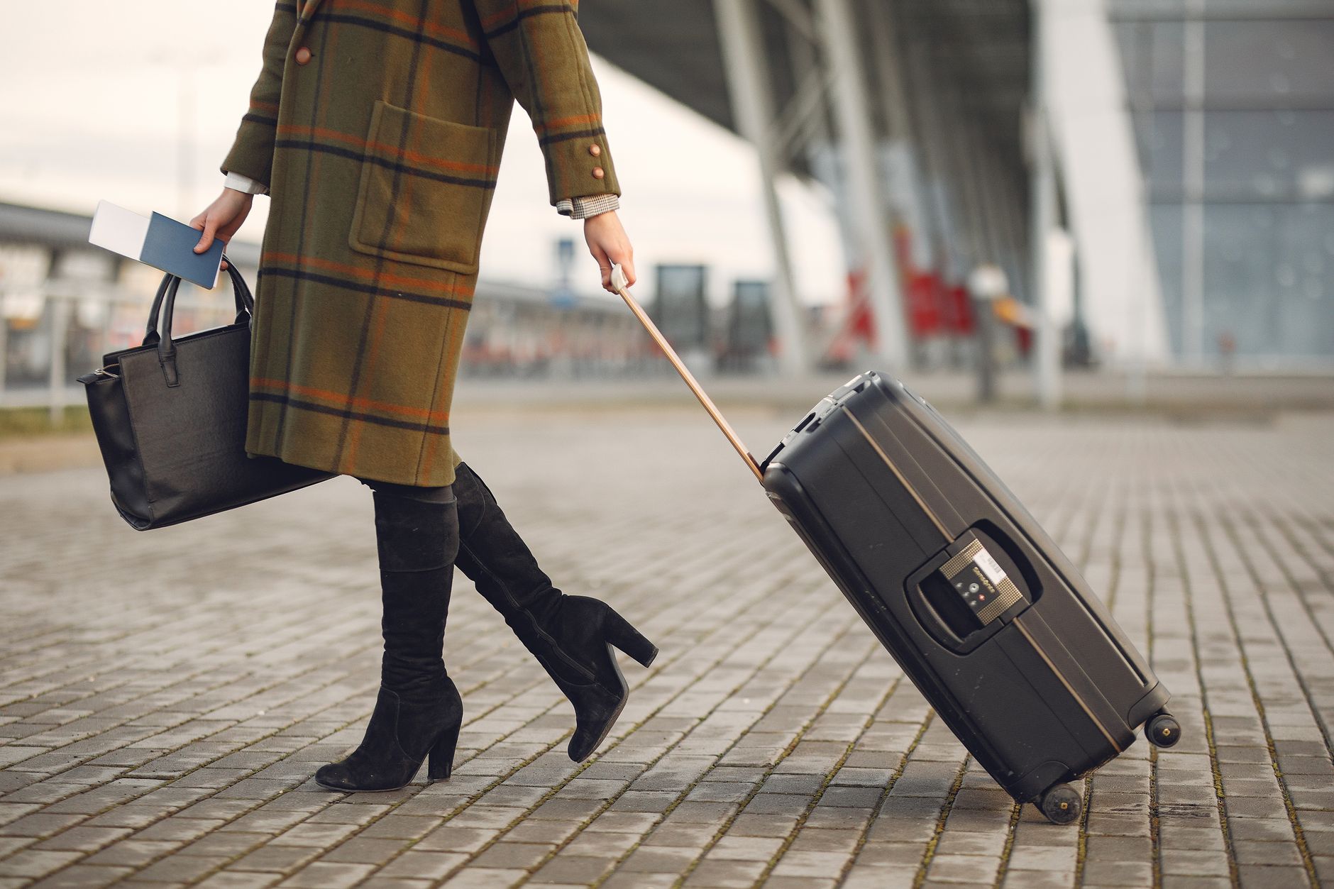Jeune voyageuse en manteau à carreaux tendance et bottes hautes avec des bagages et des documents se promenant sur le trottoir près d'un bâtiment d'aéroport contemporain pendant la saison froide