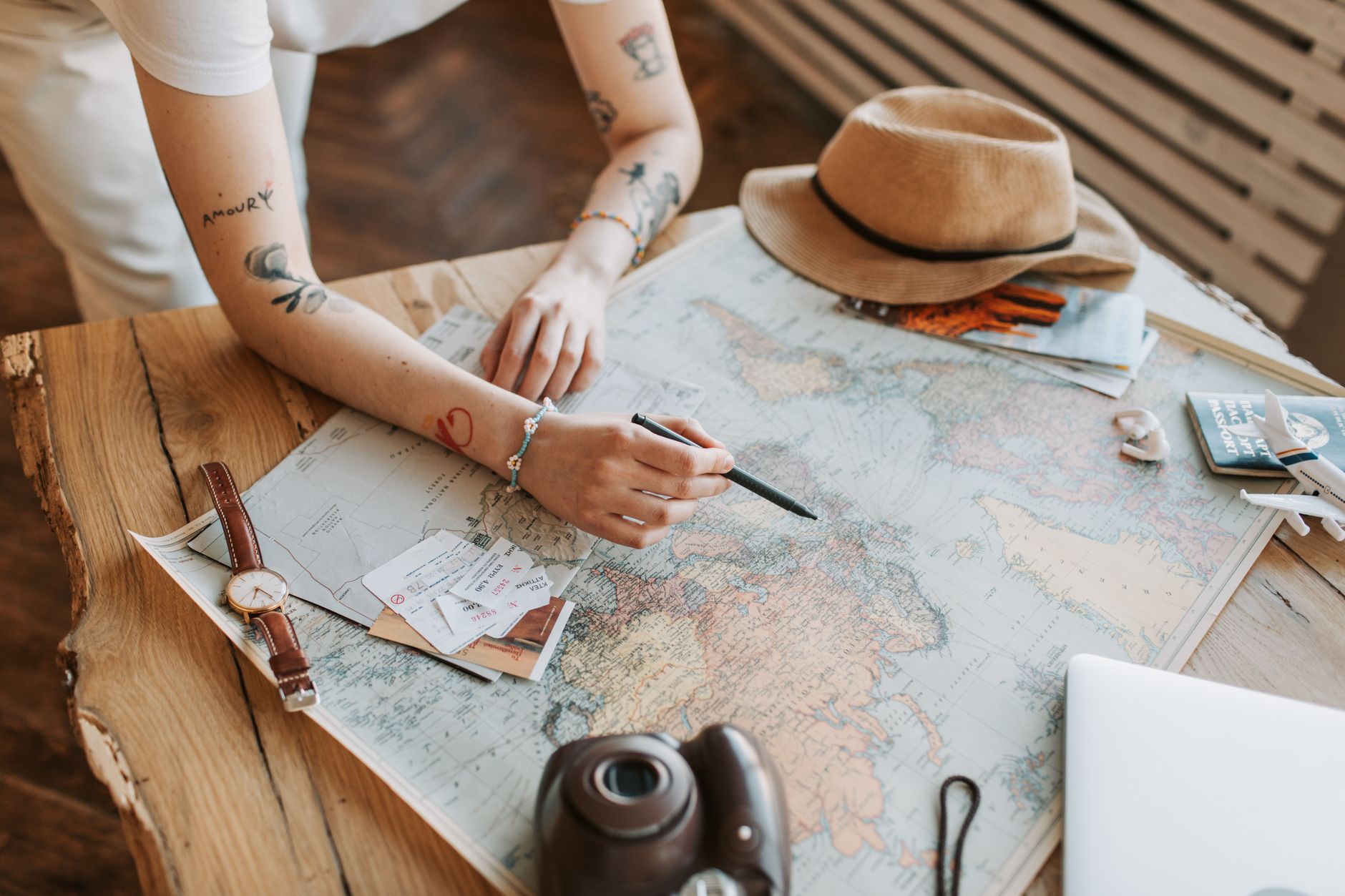 Femme tatouée planifiant un voyage avec une carte du monde, des passeports et des essentiels de voyage sur une table en bois.