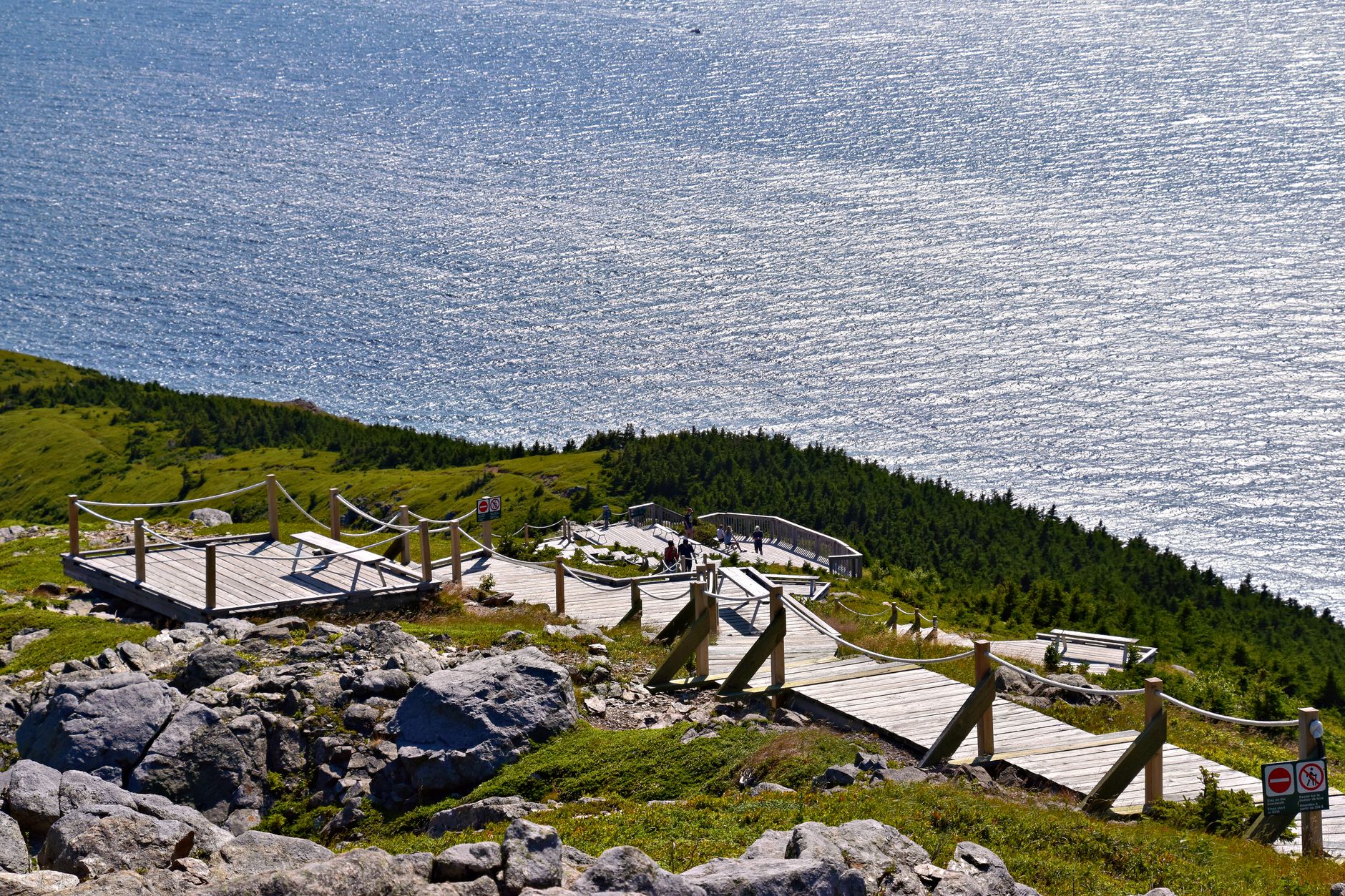 Passerelle en bois avec cordes sur une pente rocheuse et boisée, dominant une vaste mer scintillante, avec quelques randonneurs au loin.