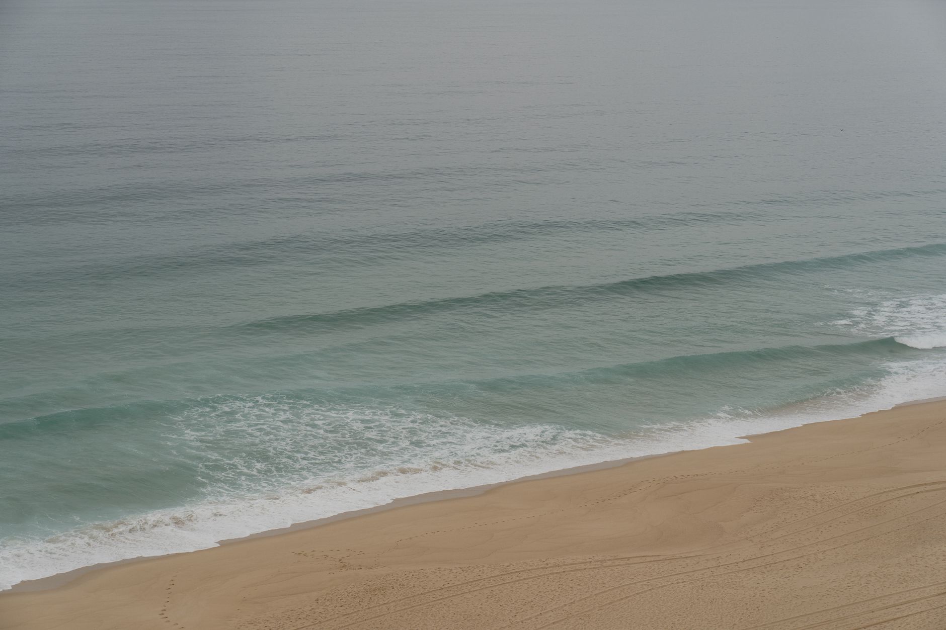 Plage de sable doré vue d’en haut, avec des vagues qui déferlent en écume blanche sur une mer vert-gris calme.